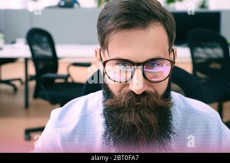Bearded man in glasses with screen reflection working on computer in IT office, sitting at desk writing code, working on a project in software Stock Photo