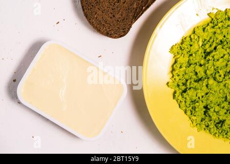 A plate of mashed avocado, cream cheese, and slices of bread on a white table in close-up. Making avocado toast. Top view, flat layout. Stock Photo