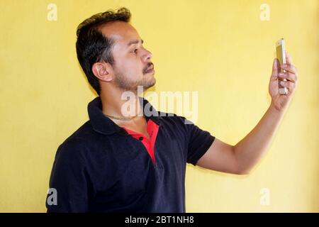 Young handsome Indian man taking selfie with mobile phone on yellow background Stock Photo