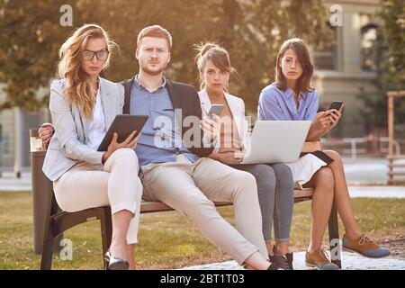 angry caucasian businesspeople sitting outdoor in park at bench, working on laptop, cell phones, tablet. Looking at camera, eye contact. business afte Stock Photo