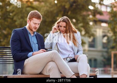 young caucasian businessman showing content on cell phone to female colleague at bench in park. serious, worried businesspeople after corona virus loc Stock Photo