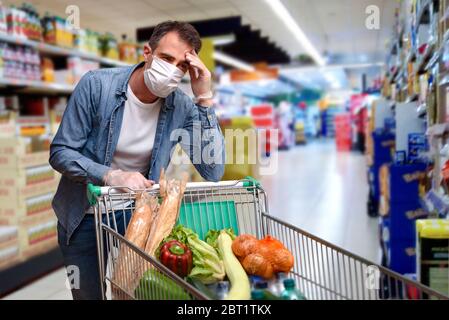 Sick man with fever leaning on a shopping cart full of food with face shields and gloves in a supermarket Stock Photo