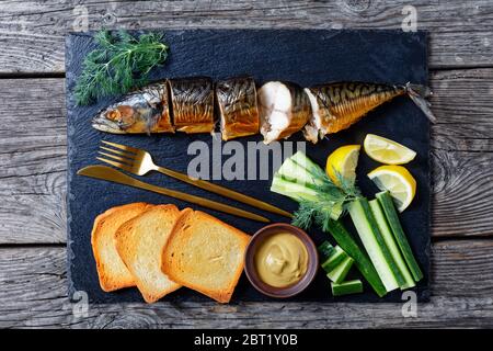 Smoked fish: smoked mackerel served on a black slate with dijon mustard, fresh cucumber, toasts, lemon wedges and cutlery on a wooden background, flat Stock Photo