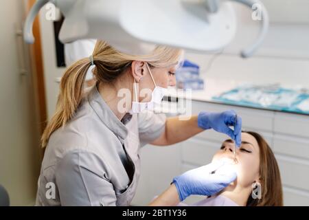 Female dentist in mask and gloves examining teeth of young female patient while working in dental clinic Stock Photo