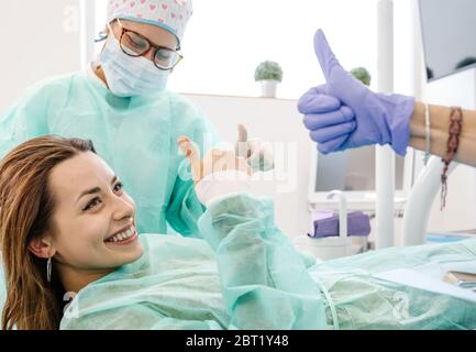 Happy female patient smiling and showing thumb up gesture with professional dentists during appointment in modern office in clinic Stock Photo