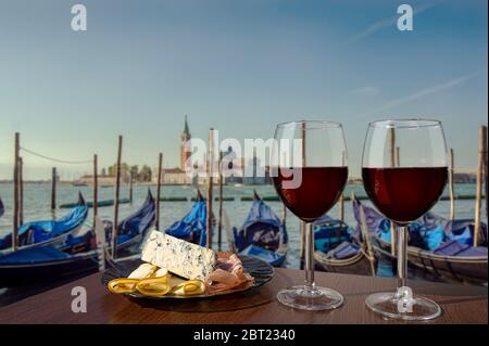 Two glasses of wine with charcuterie assortment on view of gondolas and San Giorgio Maggiore church in the background in Venice. Glass of red wine wit Stock Photo