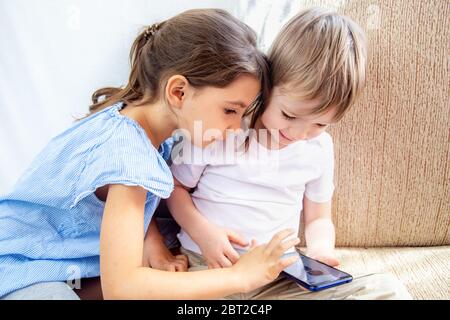 Close up siblings brother and sister playing with tablet. Kids have a good time at home. Children sitting on sofa, playing with tablet. Kids play a Stock Photo