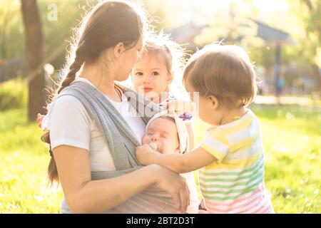 Motherhood, care, infants, summer, childhood and large families concept - Young beautiful mom with newborn baby in sling and two small children of the Stock Photo