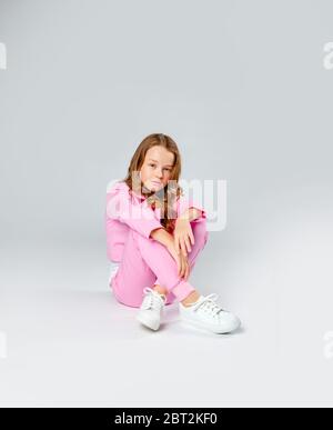 girl in a pink tracksuit and sneakers sits on a light gray background ...