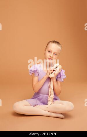 a girl in a lilac ball gown sits on the floor, legs crossed in Turkish, and gently holds pointe shoes Stock Photo