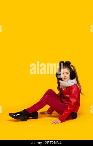 Girl in leather jacket with a scarf in red pants and fur headphones sits  on a yellow background Stock Photo