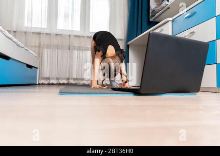a little girl in a black gymnastics leotard is doing gymnastics at home online in front of a laptop. space for text and copy space.