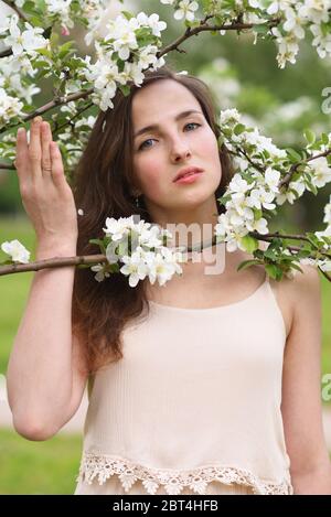 A young woman in a beige dress looks between the blossoming white branches of an Apple tree holding them with her hand Stock Photo