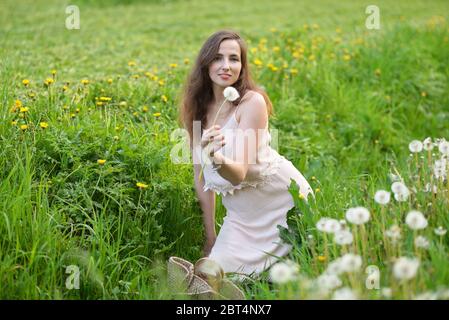 A young Russian girl in a pink dress is sitting on the green grass and holding out a dandelion Stock Photo