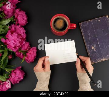 empty white postcard and two female hands, on the table a bouquet of red peonies and a ceramic cup of coffee, top view Stock Photo