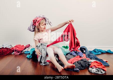 Kid playing with clothes on head. Cute Caucasian girl sorting clothes. Adorable funny child arranging organazing clothing. Messy stack of clothes thin Stock Photo