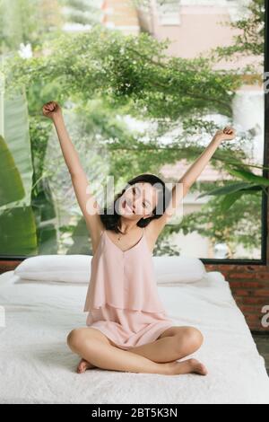 Adorable charming beautiful sweet cute funny cheerful brunette girl sitting on bed with crossed folded legs, raising hands up, Stock Photo