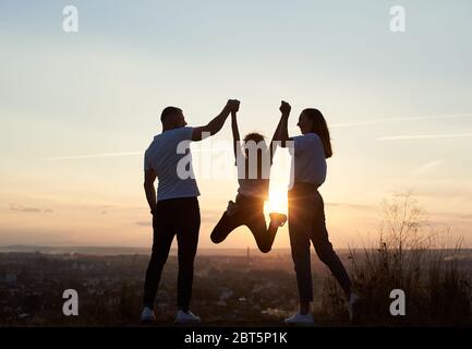 Silhouette of father, mother and daughter having fun outside the city on the hill on the sunset with a beautiful city view, man and woman holding girl by hands and she is jumping, back view Stock Photo