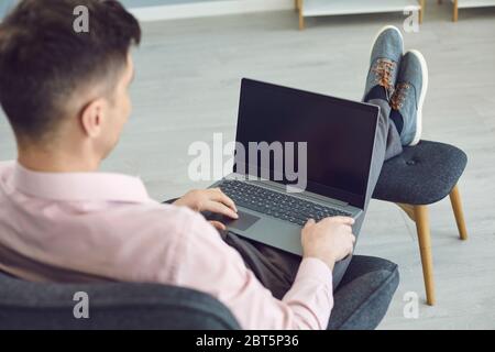 High angle of unrecognizable relaxed male sitting in armchair with legs on stool and browsing laptop with black screen at home Stock Photo