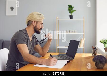 Side view of bearded young man biting glasses and thinking while reading data from laptop and making notes during remote work at home Stock Photo