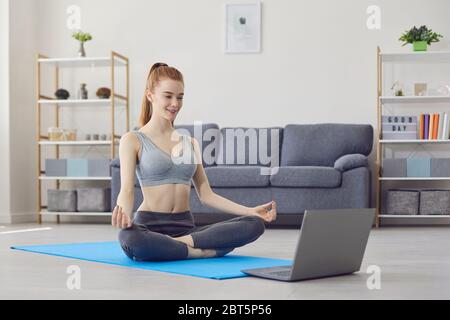 Slim young female in sportswear sitting on floor in lotus pose and meditating while watching online yoga course on laptop at home Stock Photo
