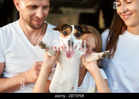Close-up shot focused on jack russell terrier held by paws a girl, father and mothers are smiling on the blured background, white family look. Dog looking to the camera Stock Photo