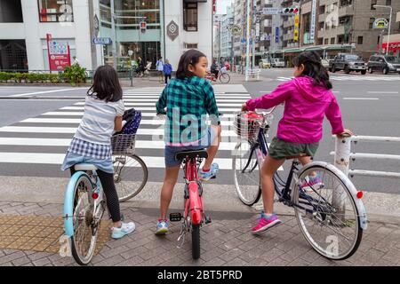 Tokyo, Japan - April 20, 2020 : three japanese girl friend children are ride bicycle and stop on pedestrian crosswalk at traffic light before crossing Stock Photo