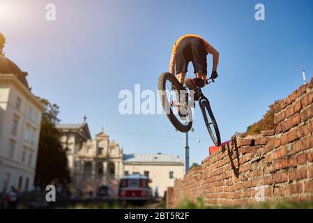 Back view of amateur cycling performing trick. Athlete in city center on a background of architecture practicing jumping on a mountain bike. Concept of sports life. Stock Photo