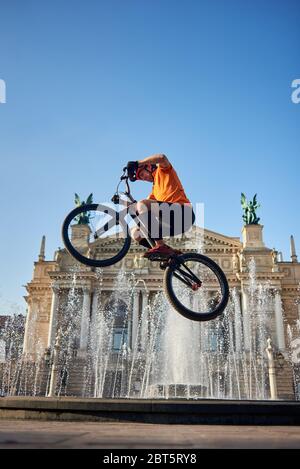View from below strong athlete jumping in air on background of fountain in city center. Guy holding bicycle handlebar and doing trick on architecture background. Concept of adrenaline. Stock Photo