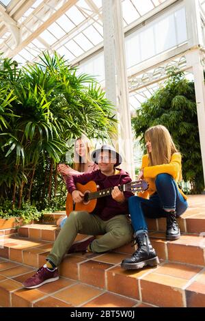 Two girls smile and enjoy themselves while a musician plays a guitar sitting on a staircase with a bright background of trees Stock Photo
