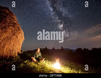 Amazing starry night in the mountains, girl is sitting on the grass near a bonfire and a rick of dry hay, meditating under bright stars, astrophotography Stock Photo