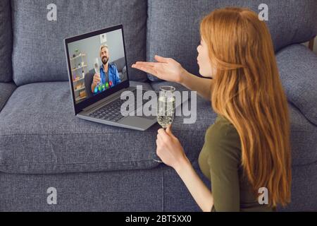 Young redhead woman with glass of champagne talking with boyfriend through video chat app on laptop while sitting near sofa at home Stock Photo