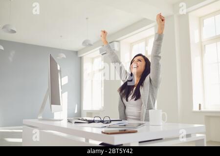 Happy successful smiling business woman raised her hands up while sitting at a table in the office. Successful transaction conclusion of an employment Stock Photo