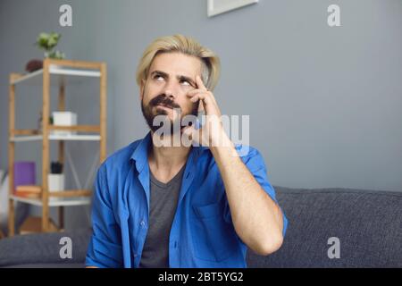 Bearded guy with dyed hair rubbing temple and looking up while sitting on sofa and pondering at home Stock Photo