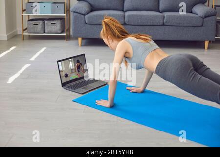 Young female doing plank exercise on mat near laptop while listening to online trainer during fitness workout at home Stock Photo