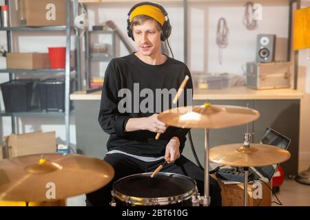 Young man in headphones, yellow beanie, black sweatshirt and jeans sitting in front of drumset while beating drum with drumsticks Stock Photo
