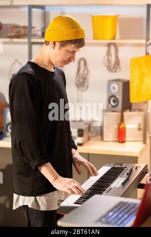 Side view of young musician in yellow beanie and black casualwear touching keys of pianoboard while recording new music in garage Stock Photo