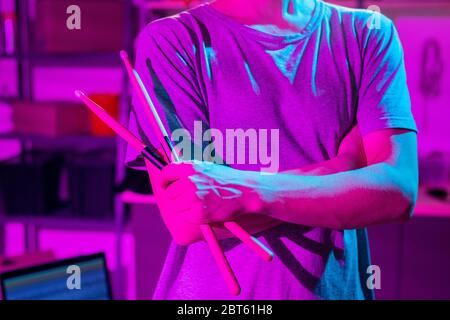 Mid section of young man in t-shirt holding drumsticks while crossing arms by chest during rehearsal with drums and cymbals in garage Stock Photo