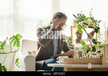 Aged bearded male professional in elegant suit and eyeglesses bending over document while looking through data while sitting in armchair Stock Photo