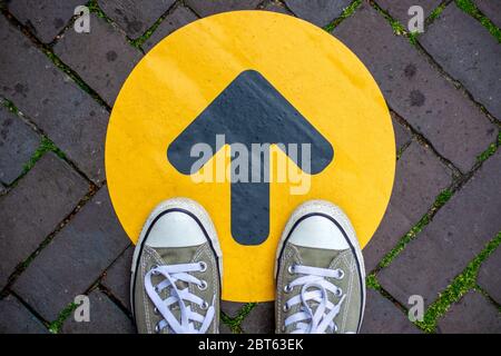 Feet wearing sneakers in front of arrow on road outdoors. Direction arrow in front of a store for the social distancing during the covid-19 pandemic. Stock Photo