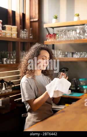 Pretty young waitress in workwear cleaning glass with white cotton napkin or towel while standing by bar counter in restaurant or cafe Stock Photo