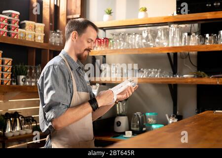 Happy young bearded waiter in apron wiping glass with napkin or towel while standing by counter among shelves with kitchenware Stock Photo