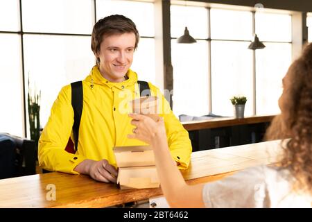 Happy young courier in yellow jacket taking two containers with ordered food and drink for clients while standing in front of waitress Stock Photo