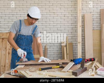 Asian carpenter with a hard hat and dustproof glasses use a sander machine to smooth plywood surfaces by abrasion with sandpaper. Morning work atmosph Stock Photo