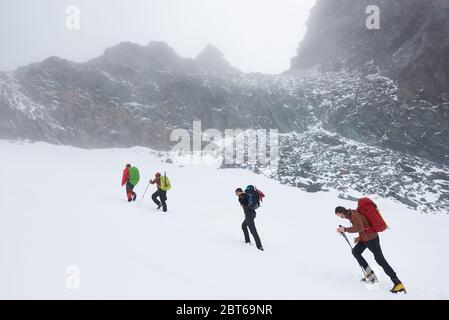 Group of brave hikers with trekking sticks and backpacks having winter hiking tour in mountains, walking through snow near big rocky hill. Concept of travelling, hiking and mountaineering. Stock Photo