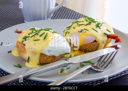 Eggs Benedict with bacon, hollandaise sauce, chives on a bun with sesame seeds. Light plate, light from the window, morning Stock Photo
