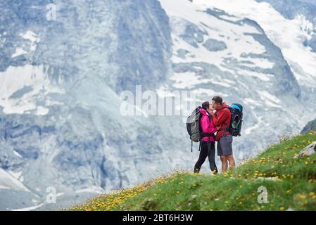 Horisontal snapshot of young couple of hikers kissing in Swiss Alps, standing on a green meadow full of yellow flowers, behind them magnificent snowy mountain, concept of active lifestyle Stock Photo