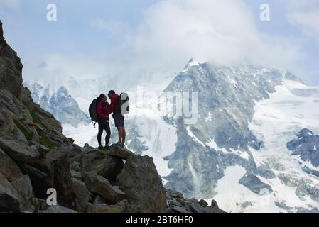 Happy couple hikers with backpacks. Beautiful mountains scenery on background. Trekking, tourists reaching peak together. Wild nature with amazing views. Sport tourism in Alps. Stock Photo