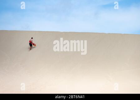 Kangaroo Island, Australia - March 9th, 2020: A young man climbing a sand dune for sand surfing. Stock Photo