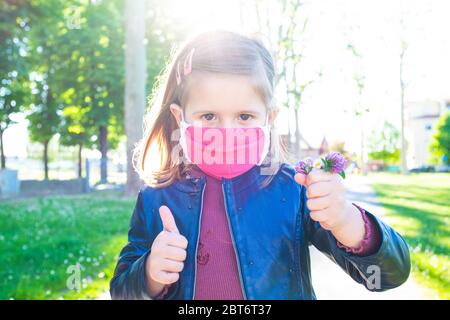 Cute little girl shows thumb up, wearing protective mask and holding bunch of wildflowers - Coronavirus time, Everything Will be fine - Optimism and c Stock Photo